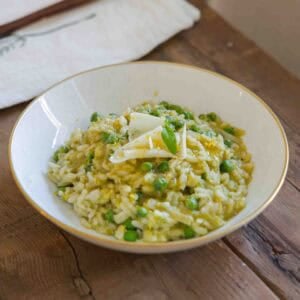 A white bowl with a gold rim holds creamy risotto mixed with green peas and topped with cheese shavings and herbs. The bowl sits on a rustic wooden table next to a white napkin.